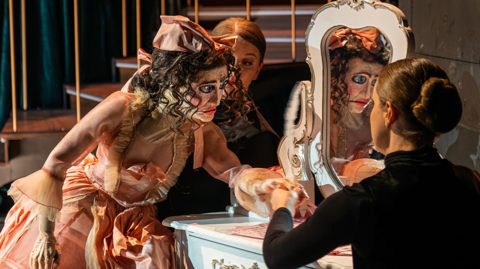 Eine Frau mit und eine frau ohne Maske auf einer Bühne vor einem Spiegel. 