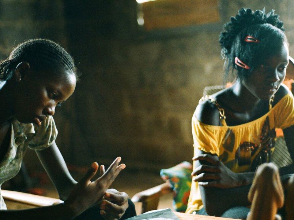 Zwei schwarze Mädchen sitzen in einem dunklen Raum. Eines der beiden schaut auf die eigenen Finger, das andere blickt ins Leere.
