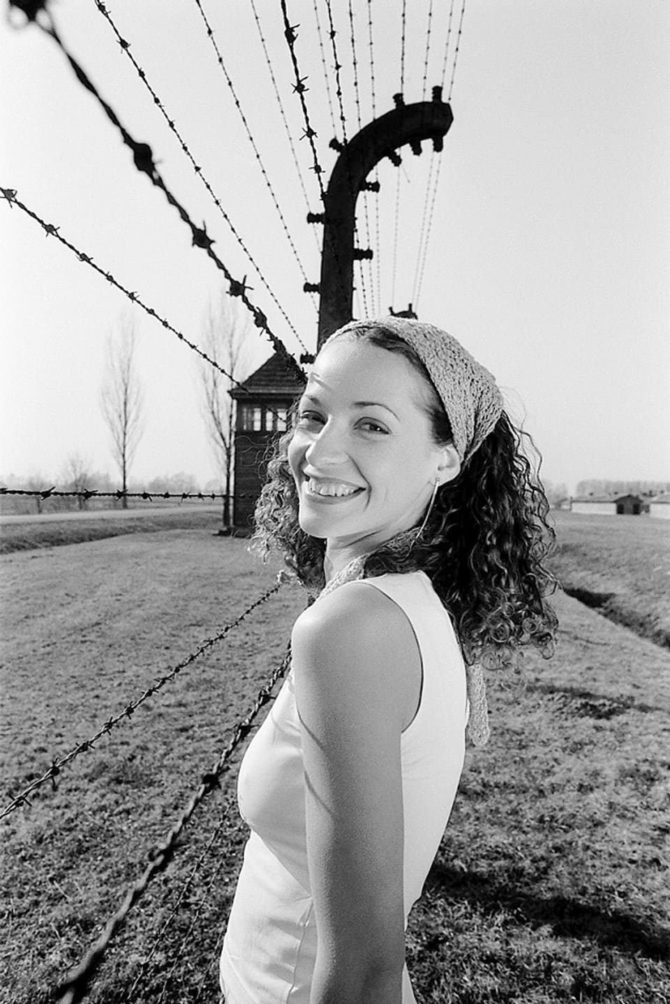 Eine Frau steht lächelnd vor einem Gitterdraht eines Konzentrationslagers. 