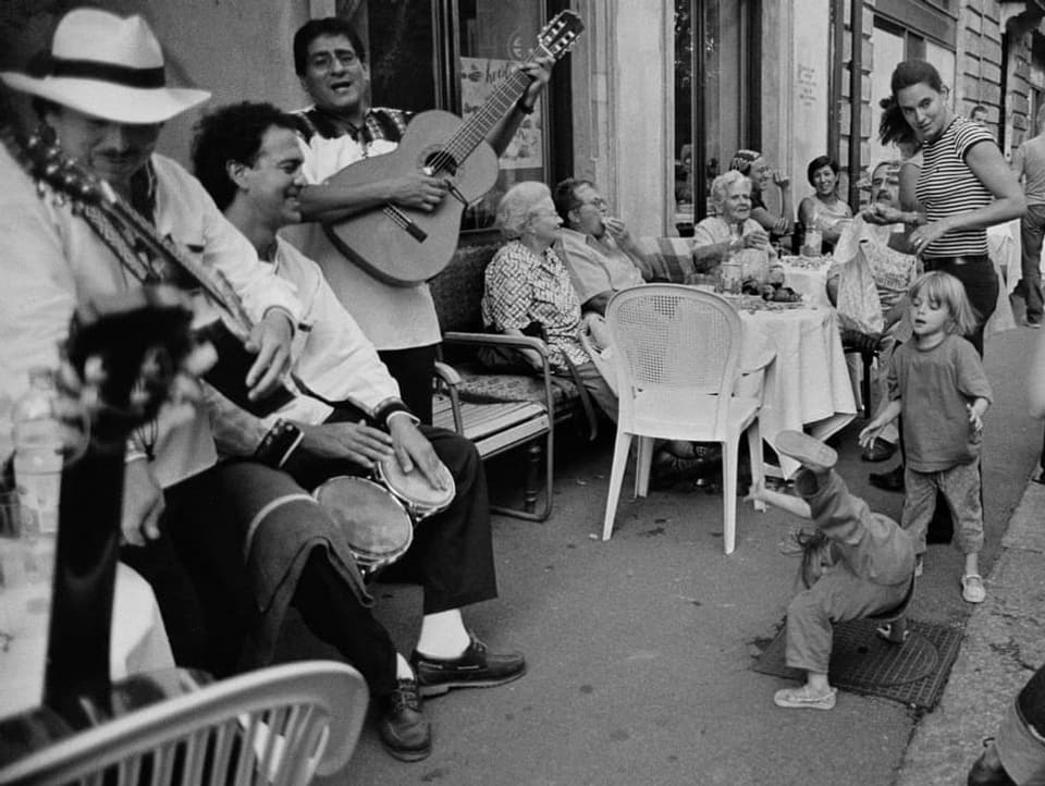 Schwarzweiss-Foto:  Menschen Musizieren vor einem Restaurant, Kinder tanzen.