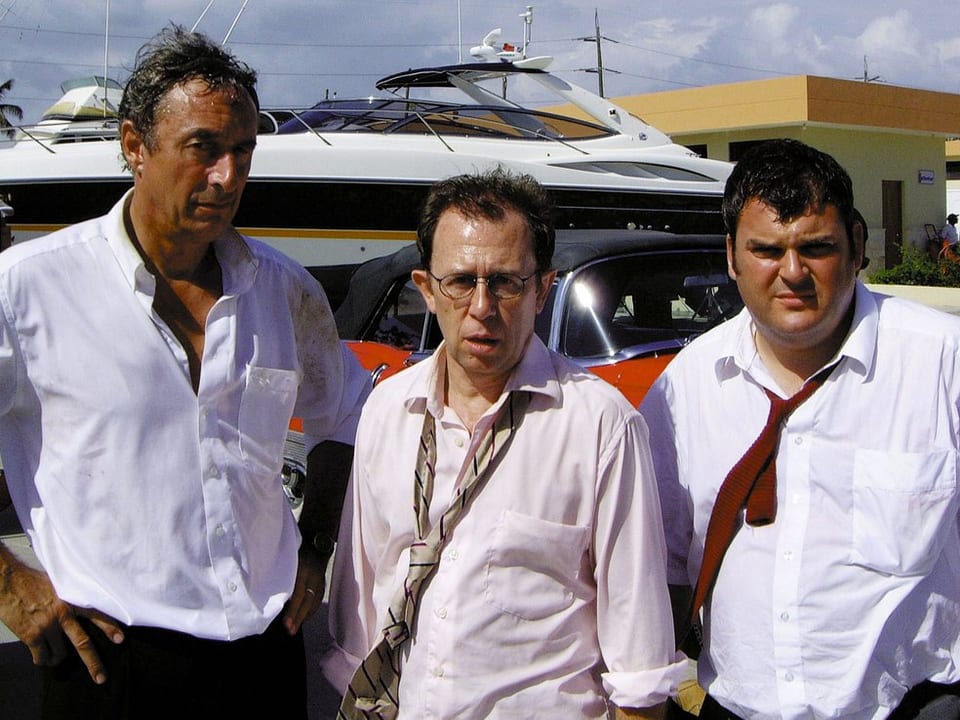 Die drei Männer stehen mit aufgeknöpftem Hemd vor einer Yacht.
