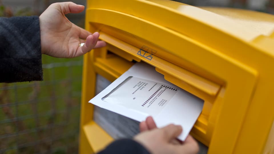 Eine Person wirft ein Wahlcouvert in einen gelben Briefkasten der Post.