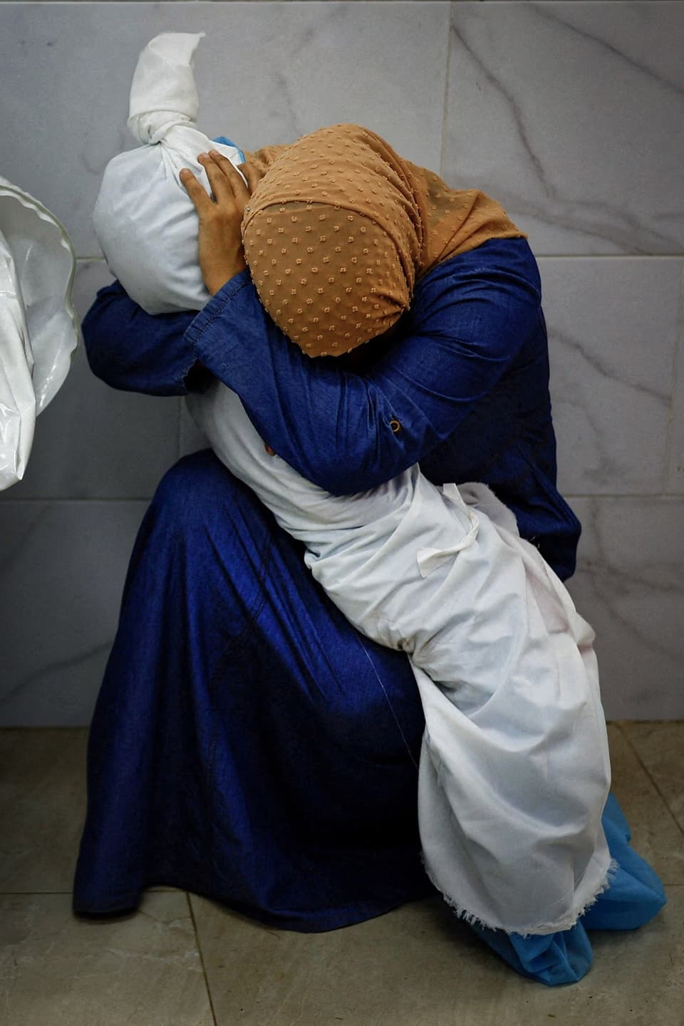 Frau in blauem Kleid und mit orangem Kopftuch, umarmt in Laken eingewickelte Leiche