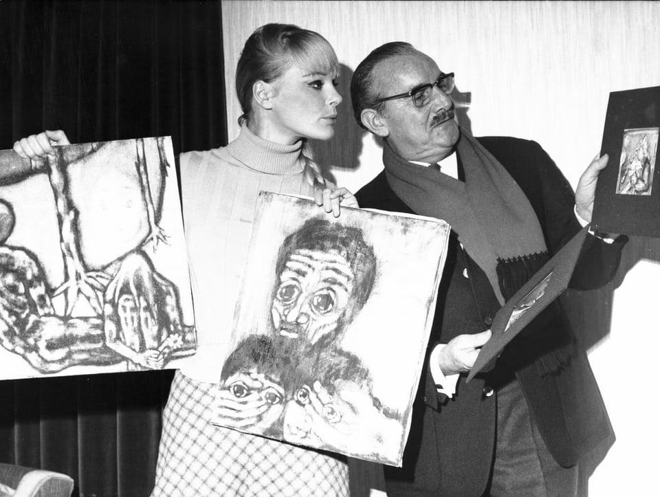 Elke Sommer mit Zopf hält zwei Gemälde in der Hand und schaut auf das Bild eines Mannes neben ihr.