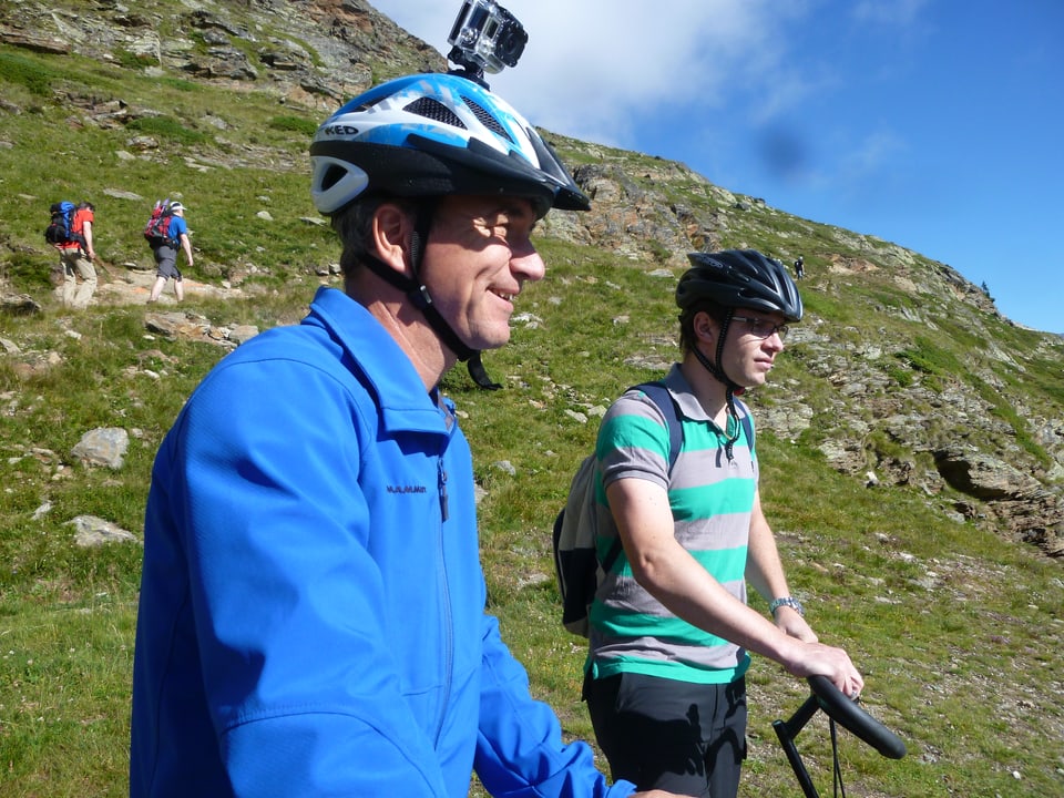 Zwei Männer stehen mit ihren Trottinetten in einer Berglandschaft. Einer hat eine Kamera an seinem Helm.