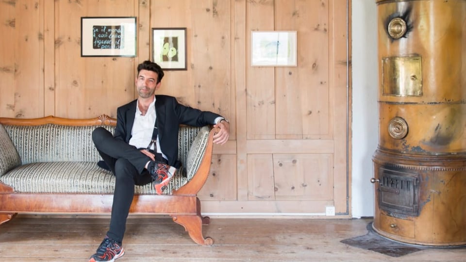 Schlanker Mann mit dunklem Haar auf altem Sofa, ein Bein angewinkelt. In Raum aus Holz, rechts Kamin.