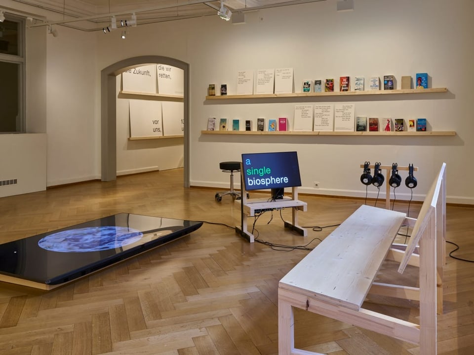 Blick in einen Ausstellungsraum mit Büchern auf einem Regal, mehreren Bildschirmen und einer Audio-Installation.