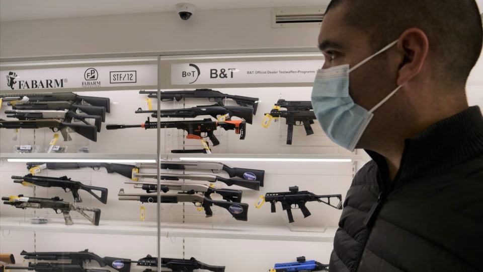 Mann mit Maske steht vor einem Glaskasten mit ausgestellten Gewehren