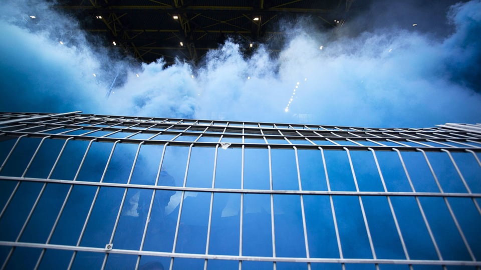 Ein Stadiongitter: dahinter sind Fans und blauer Rauch.