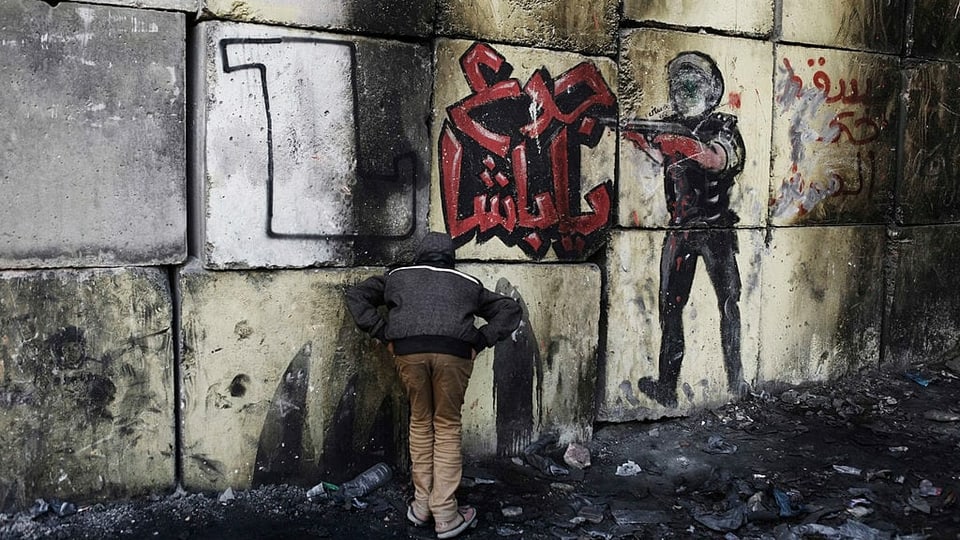 Ein Junge steht vor einer Wand aus hellgrauen Betonblöcken, worauf ein Graffiti gemalt sind.