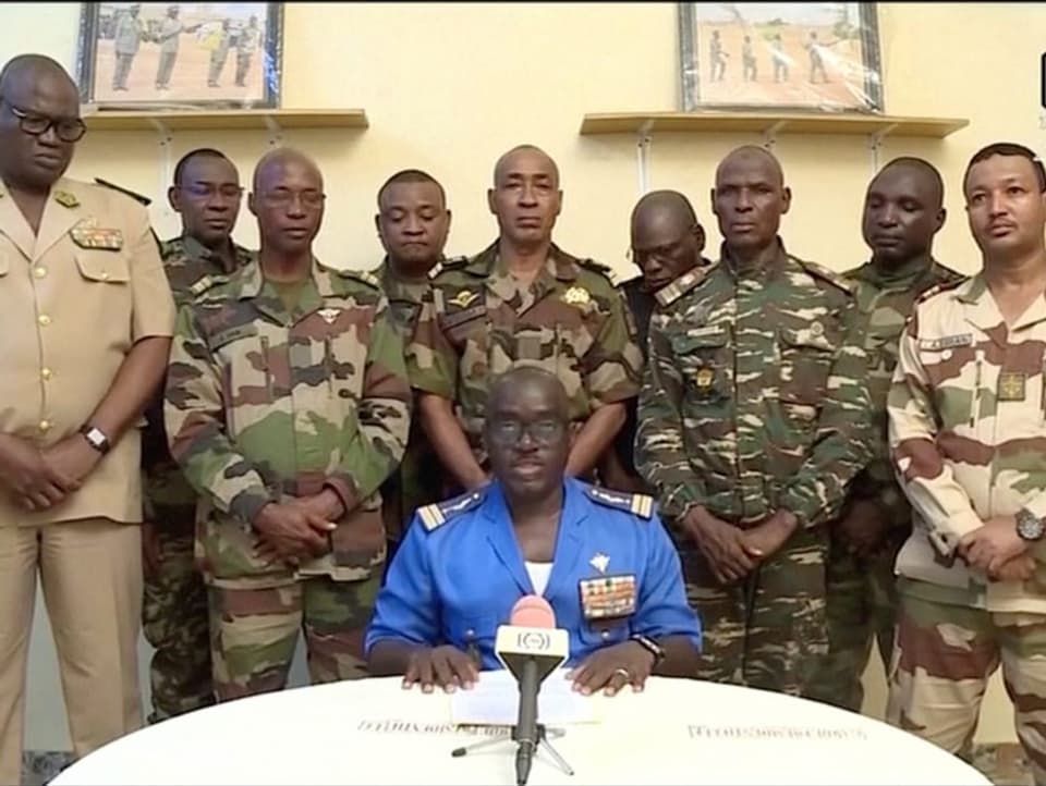 Ein Mann im blauen Anzug sitzt vor einem Mikrofon. Hinter ihm stehen mehrere Militärangehörige in Tarnanzügen.