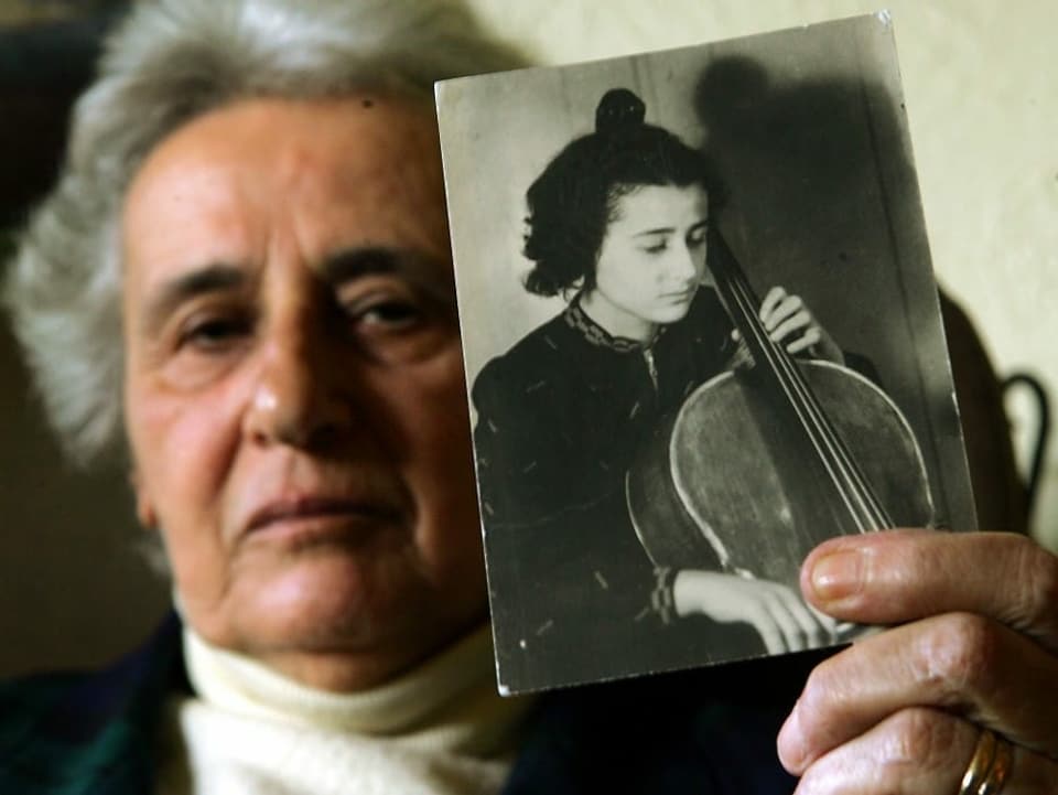 Eine ältere Frau hält ein Schwarzweissbild einer jungen Cellistin in die Kamera.