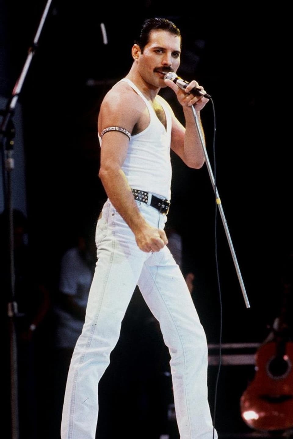 Mercury steht auf der Bühne mit schnauz, Trägershirt und Mikrofon in der linken Hand.