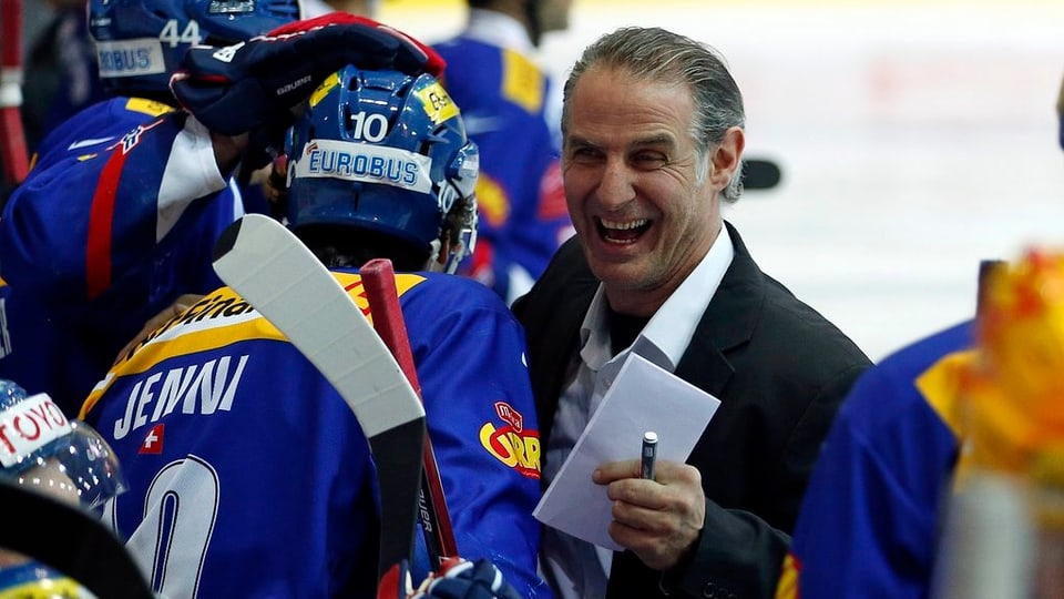 Links Eishockeyspieler in blauem Dress und blauem Helm, rechts Trainer Felix Hollenstein, kurze, graue Haare, ein Block in der Hand.