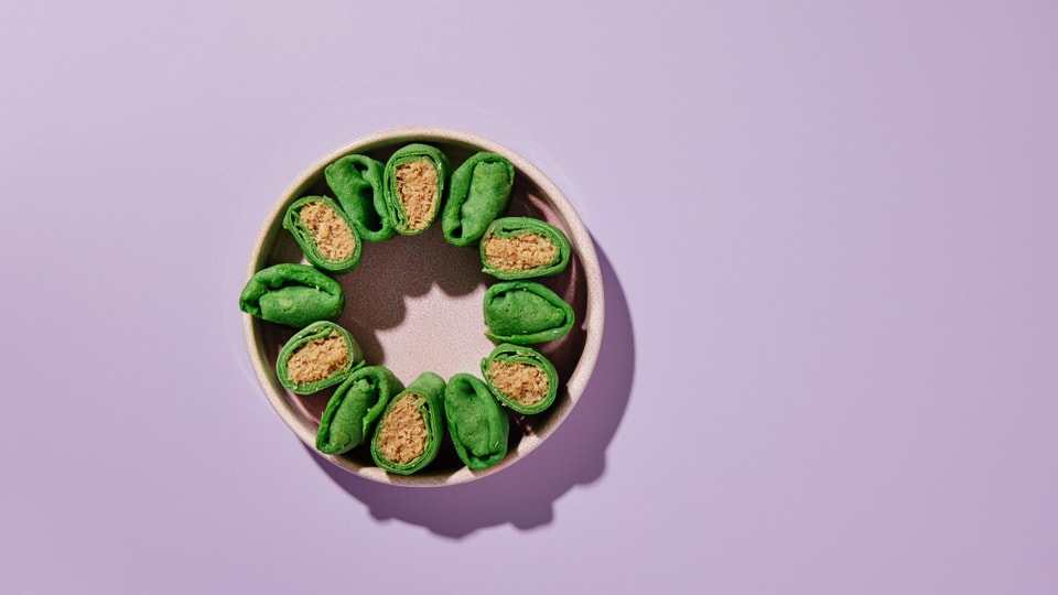 Grüne Teigrollen mit beiger Füllung, kreisförmig angeordnet in einem Teller vor fliederfarbenem Hintergrund