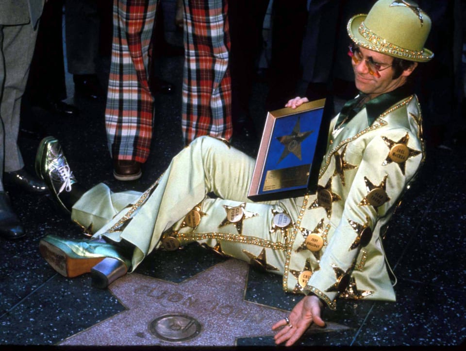 Ein Mann in ausgefallenem Glitzerkostüm sitzt auf einem Stern, der in den Boden eingelassen ist und seinen Namen trägt.