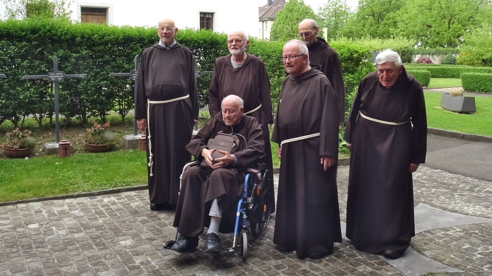 Gruppe von Mönchen in Roben, einer im Rollstuhl, im Klostergarten.