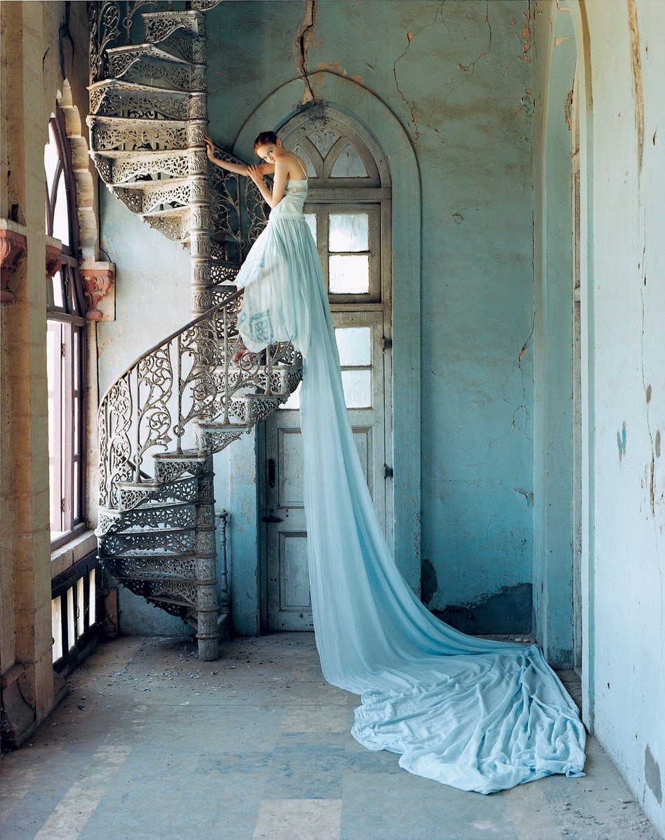 Eine Frau steht auf einer offenen Wendeltreppe ihr Kleid ist aus blauem, fliessendem Stoff. Das Kleid fällt die Wendeltreppe hinunter und reicht bis an den Boden. 