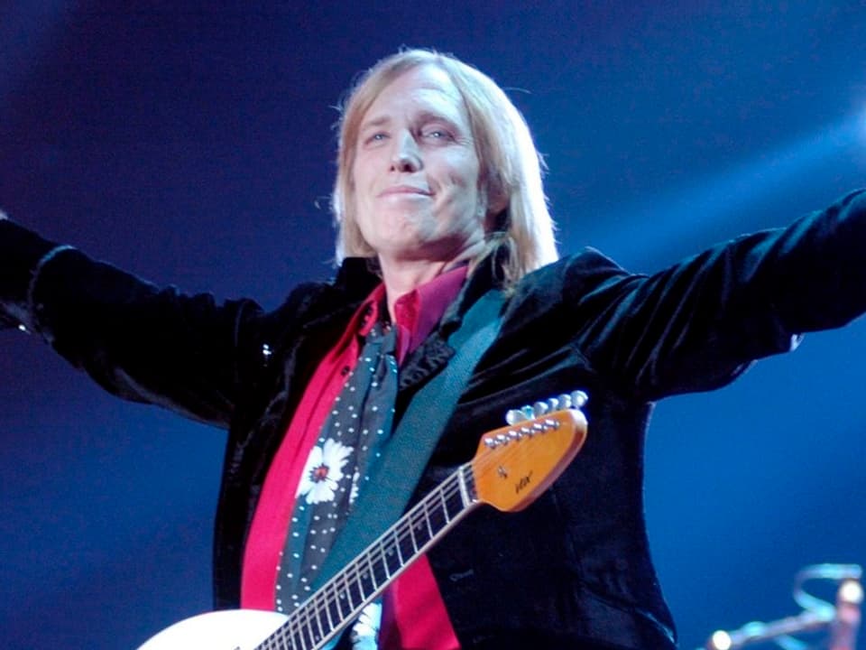 Tom Petty im schwarzen Anzug breitet die Arme aus. Er steht auf einer Bühne und hat sich eine Gitarre um den Hals gehängt.