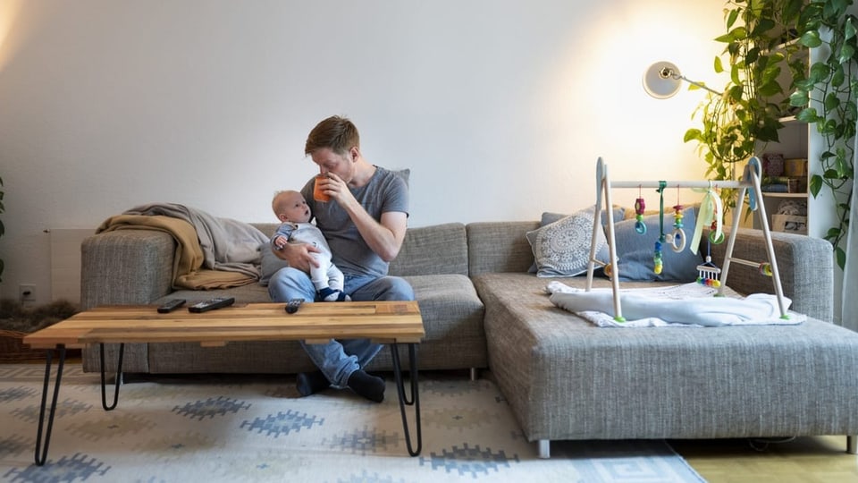 Ein Mann sitzt in seinem Wohnzimmer auf der Couch und hält sein Baby.