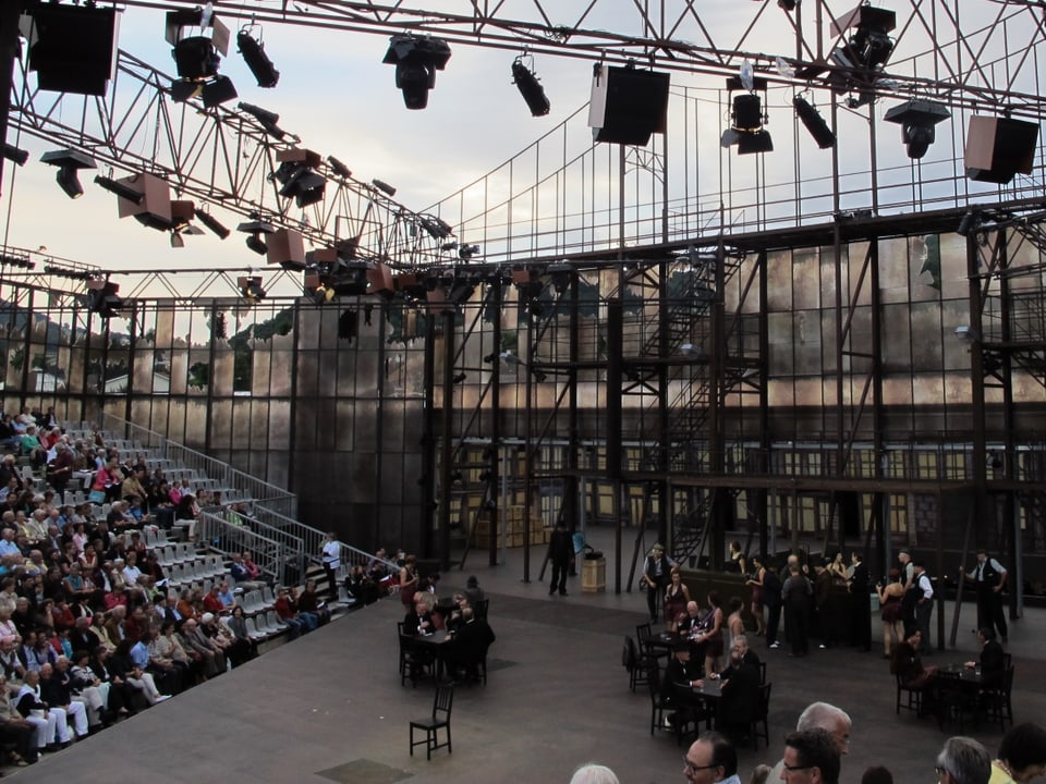 Blick in die Verdi-Arena, ein Gerüst aus Stahl, Schauspieler auf der Bühne, Publikum im Zuschauerraum.