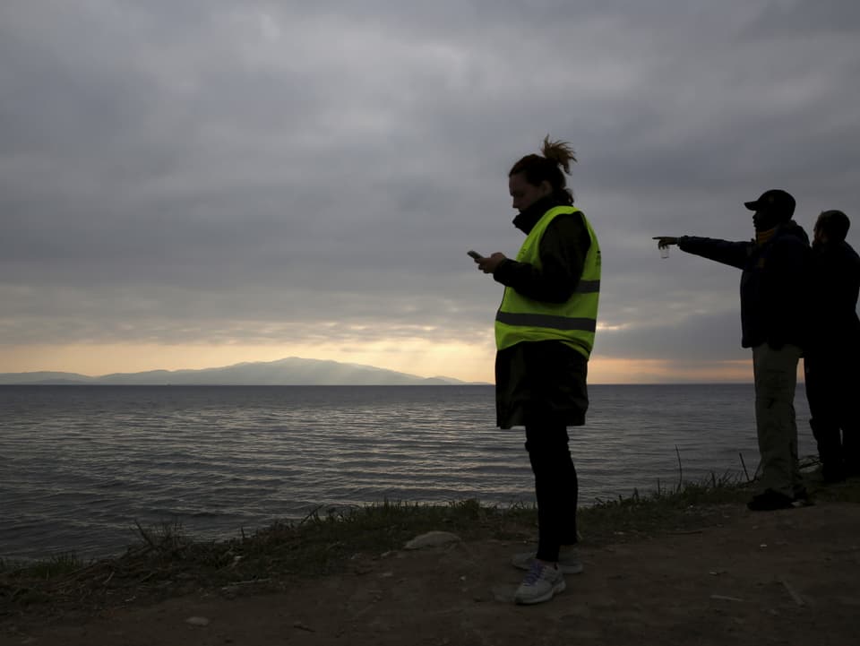 Drei Personen stehen am Strand, eine zeigt aufs Meer hinaus, eine andere – sie trägt eine gelbe Signalweste – schaut auf ihr Handy, das sie in der Hand hält.