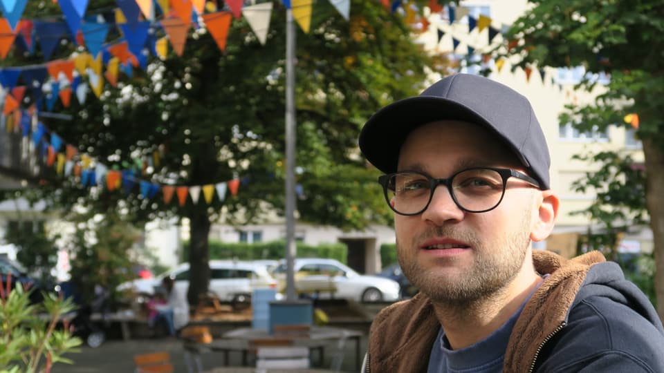 Ein Mann mit Brille und Dreitagebart im rechten Bildrand, links Tische und Stühle, sowie farbige Fähnchen.