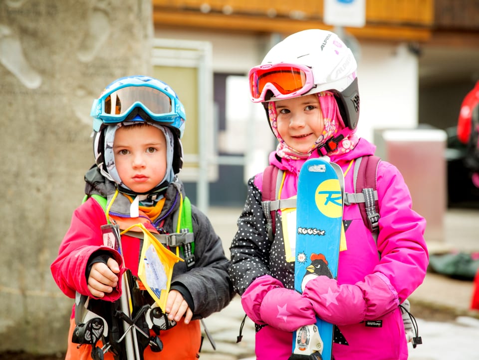 Zwei Kinder mit Helm und Skibrille halten ihre Skier in den Händen.