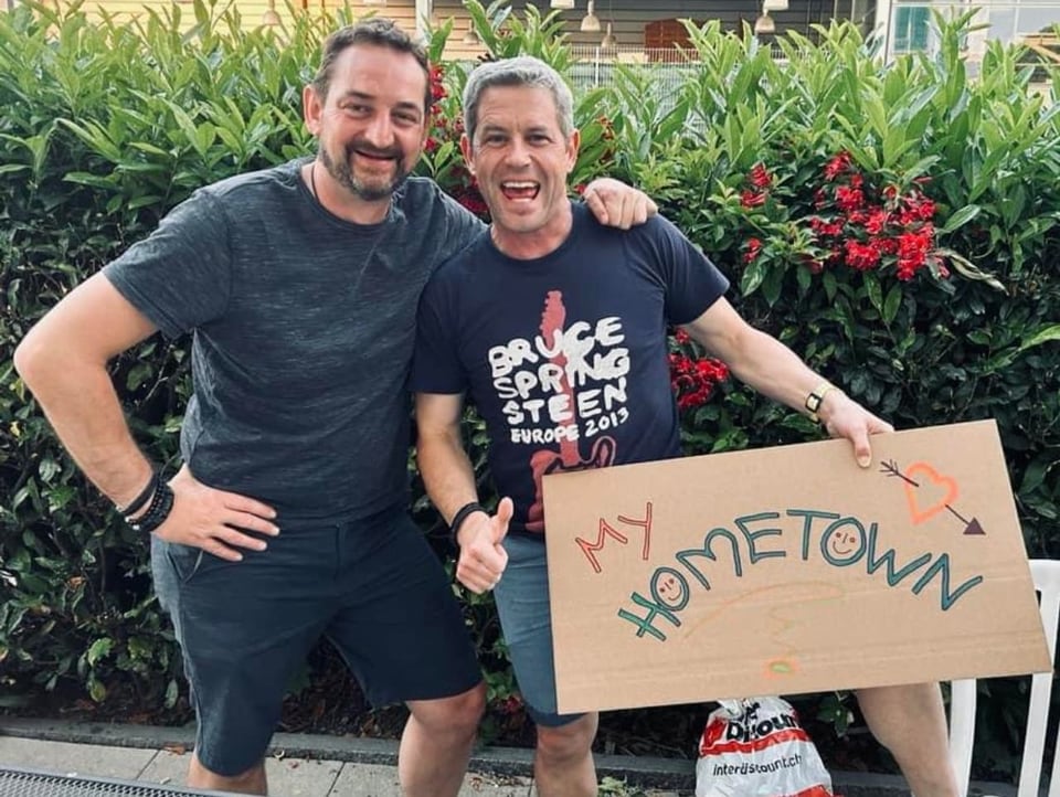 Zwei Männer posierend lachend. Der rechte Mann hält ein «My Hometown»-Plakat in der linken Hand.