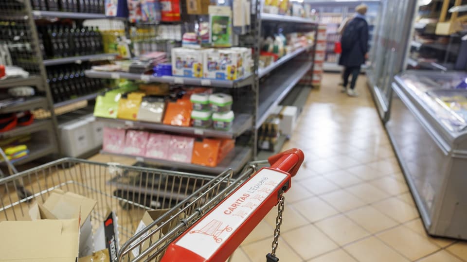 Ein Einkaufswagen mit der Aufschrift «Caritas» steht in einem Supermarkt. Eine Person ist im Hintergrund zu erkennen.