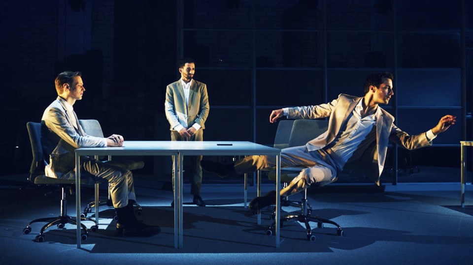 Zwei Männer in Anzügen sitzen an einem Tisch. Dahinter steht ein Dritter, ebenfalls im Anzug.