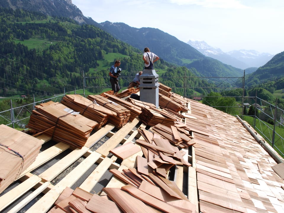 Hausdach, das mit Holzschindeln neu eingedeckt wird, dahinter eine wunderschöne Aussicht auf die Berge. 