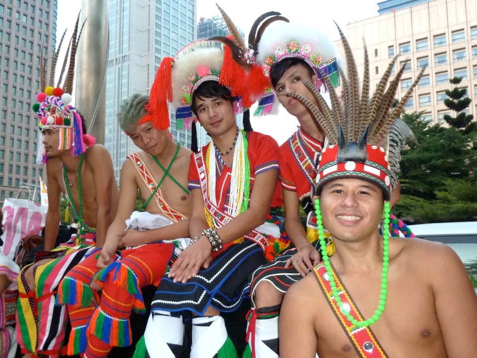 Fünf junge Männer mit nacktem Oberkörper und traditionellem Kopfschmuck sitzen vor modernen Hochhäusern.