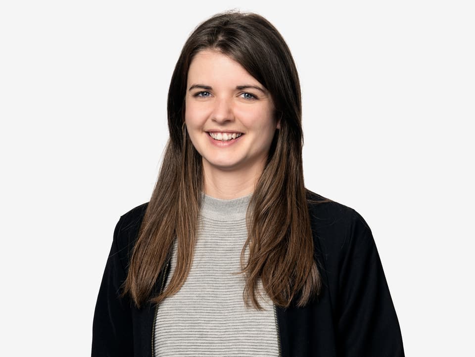 Portraitbild einer jungen Frau mit einer dunklen Bluse und grau gestreiften Pullover. Sie lächelt.