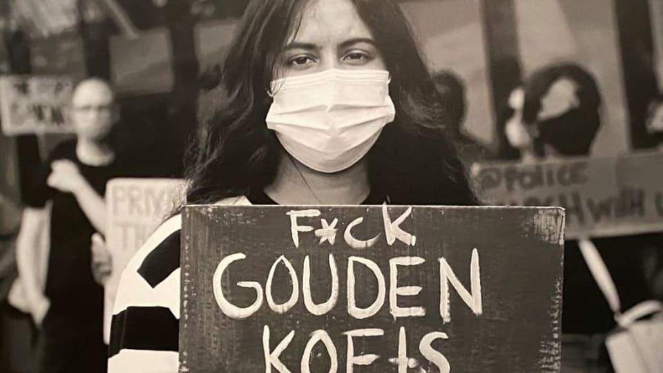 Eine junge Frau mit Maske und handgeschriebenem Plakat, auf dfem steht: «F*ck GOUDEN KEOTS»