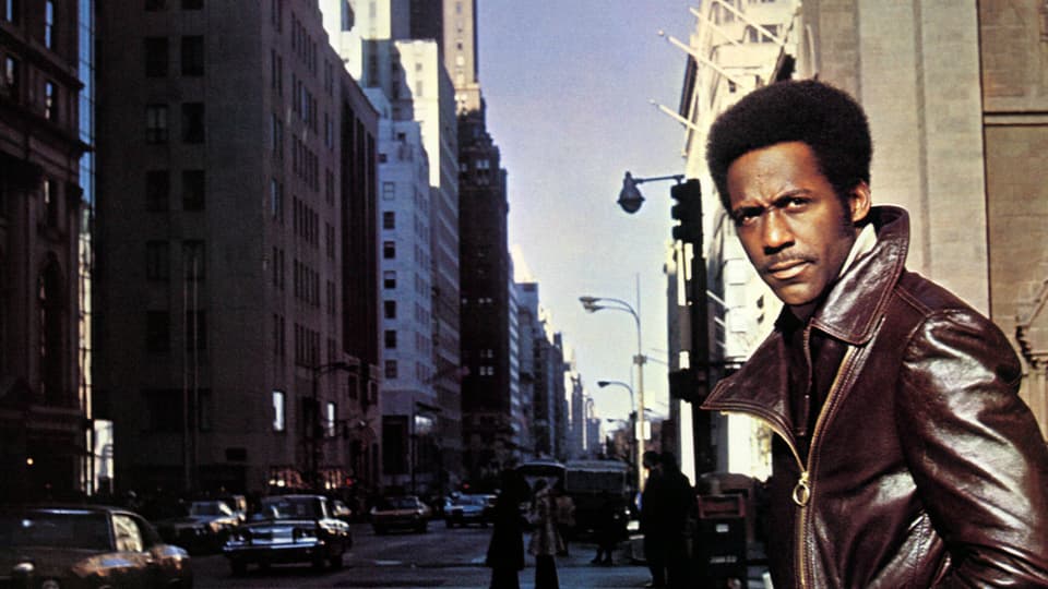 Ein schwarzer Mann mit Lederjacke steht an einer Strasse. Im Hintergrund: New York