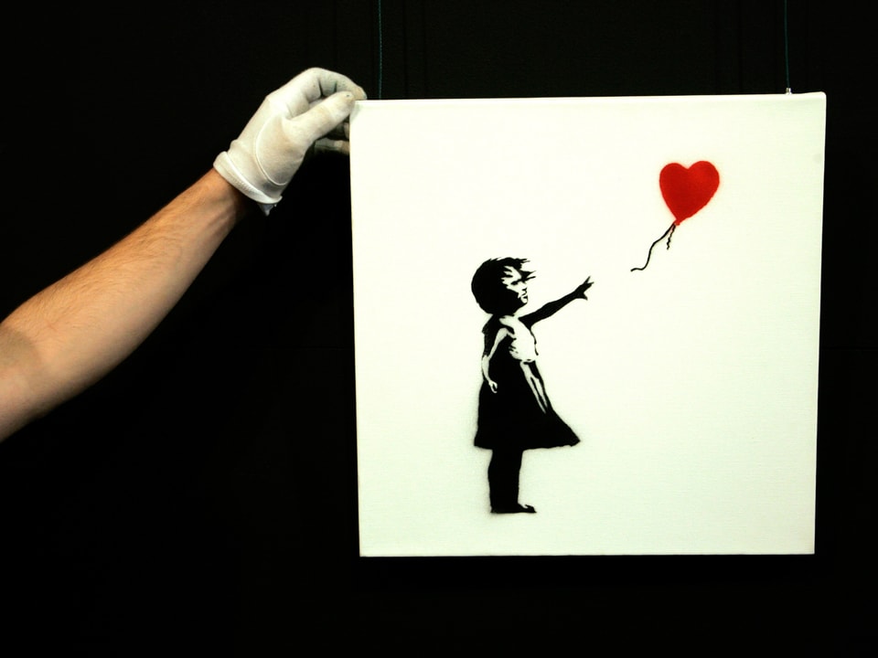 Eine Hand mit weissem Handschuh hält ein quadratisches Bild, auf dem ein gezeichnetes Mädchen mit einem roten Herzballon zu sehen ist.