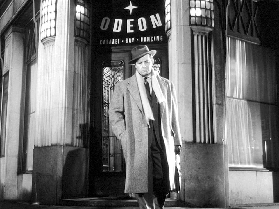 Ein Mann mit Mantel, Hut und Aktentasche steht auf der Strasse vor dem Eingang des Café Odeon.