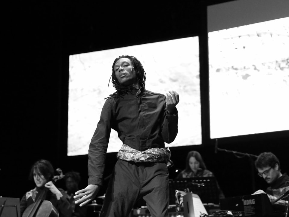 Mann mit dunkler Hautfarbe und Dreadlocks steht auf der Bühne in der Mitte in tanzender Pose, hinter ihm ein kleines Streichorchester.