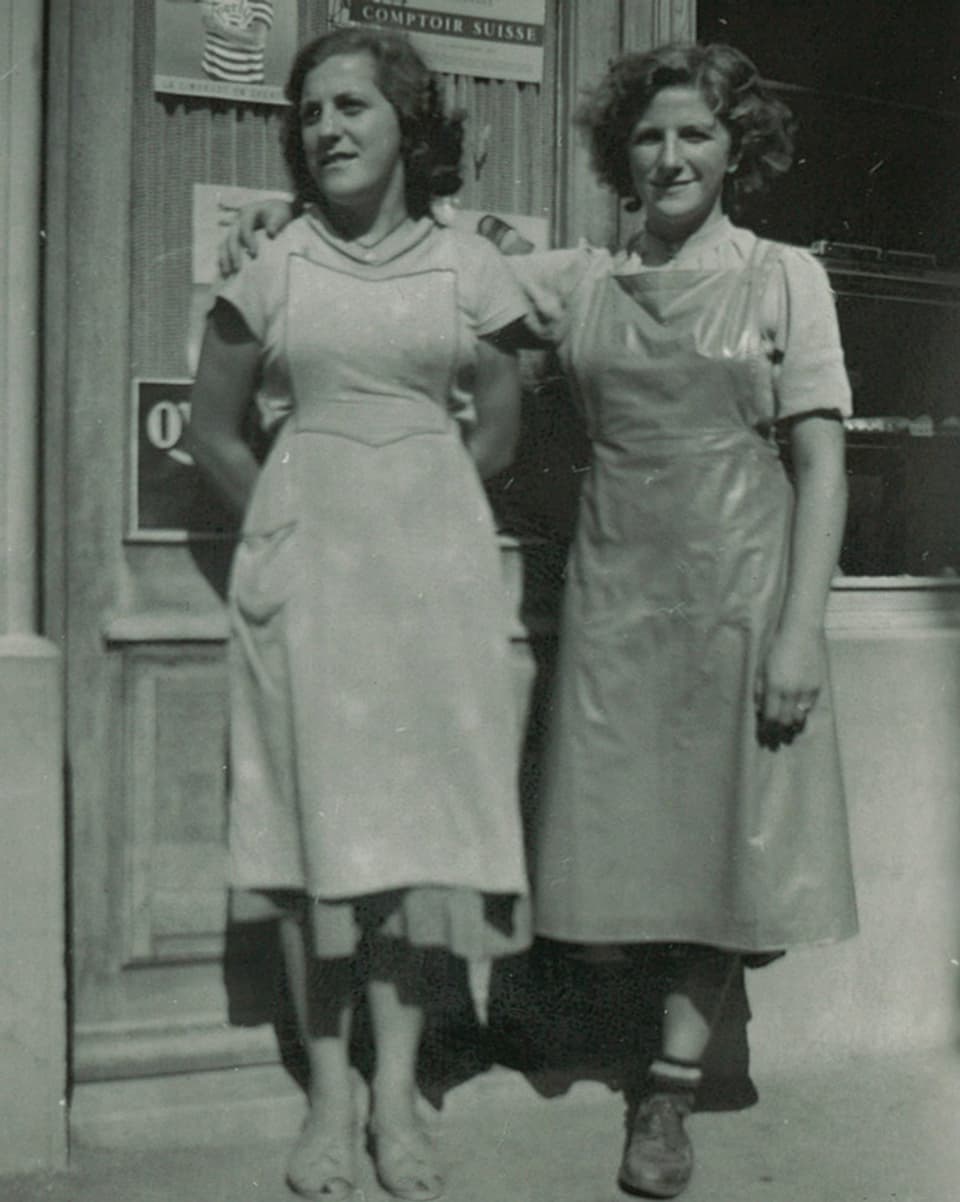 Zwei Frauen mit Schürzen stehen nebeneinander vor einer Bäckerei.
