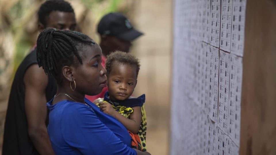 Frau mit Kind auf dem Arm sieht sich Wahllisten an Wand an.