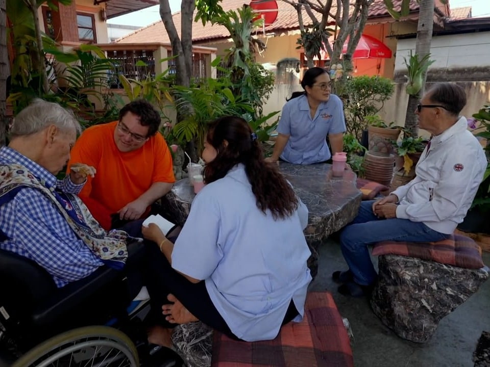 Ein älterer Herr im Rollstuhl, eine Betreuerin, ein Mann in orangen Tshirt und zwei weitere Personen sitzen am Tisch.