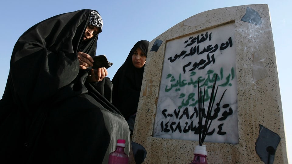 Zwei Frauen im Tschador sitzen neben einem Grabstein. Davor stehen Flaschen mit Räucherstäbchen.