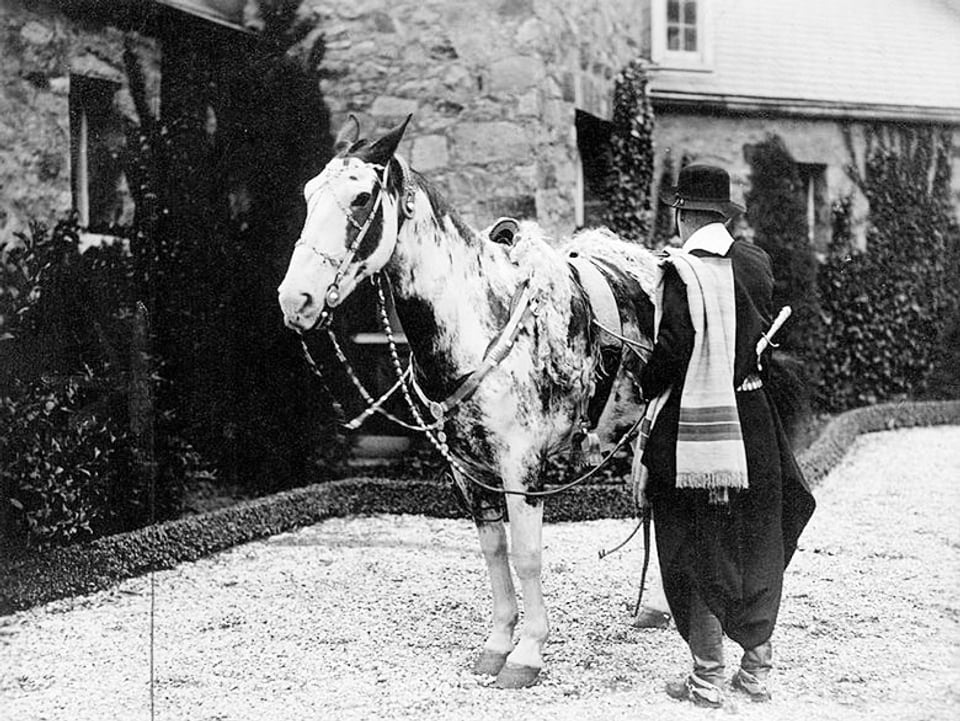 Die Archivaufnahme zeigt Aimé Félix Tschiffely, der neben seinem Pferd Mancha steht, mit dem Rücken zur Kamera.