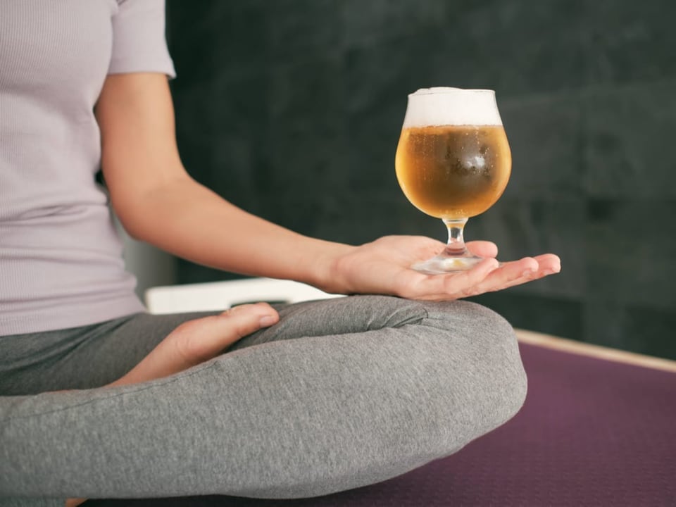 Frau sitzt im Lotussitz auf Yogamatte und balanciert ein Bier