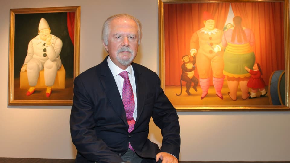 Ein alter Mann sitzt in Anzug vor zwei Gemälden.