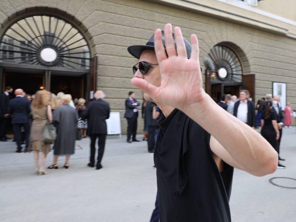 Ein Mann in schwarzem ärmelfreien Shirt, mit Sonnenbrille und Hut streckt abwehrend die Hand in Richtung Kamera