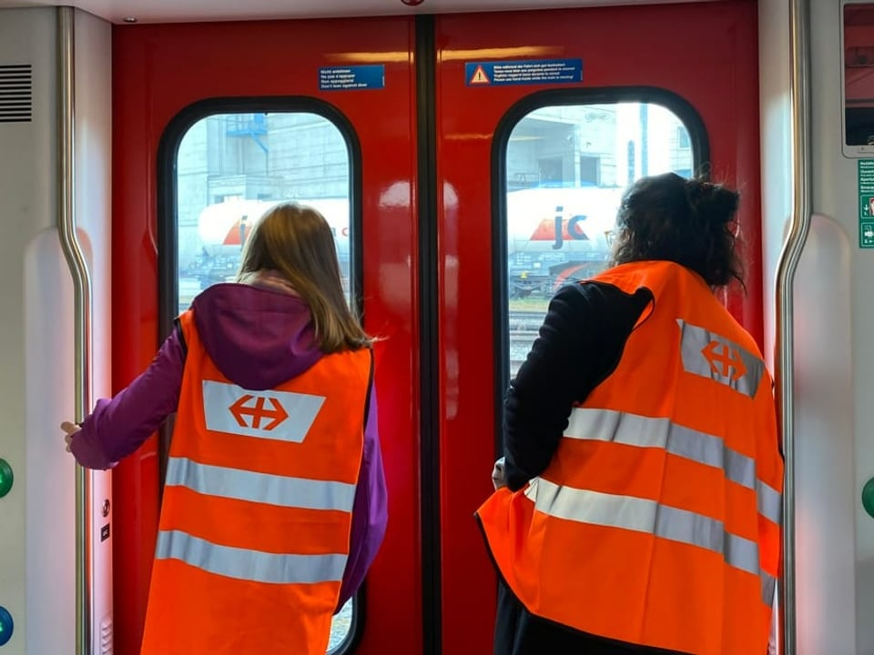 Die beiden Frauen in Warnwesten stehen vor der geschlossenen Zugtür und schauen durch die Fenster.