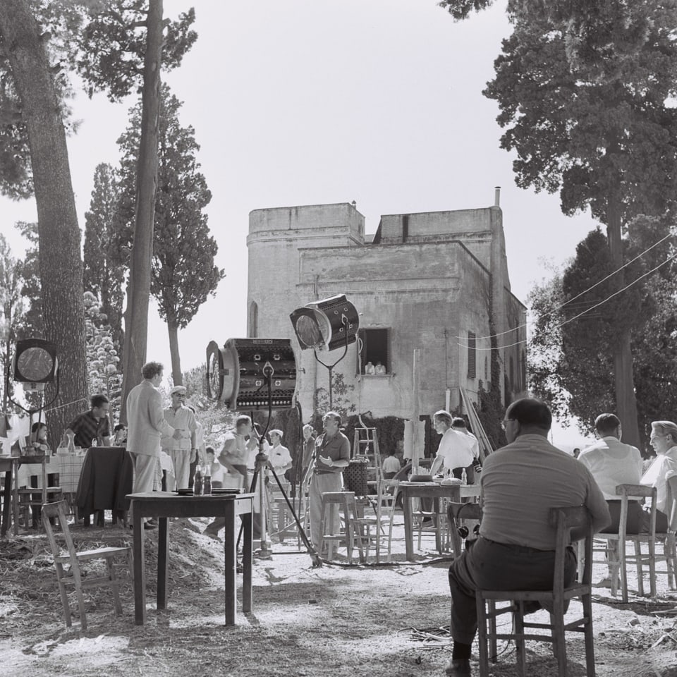 Filmset in Italien. Ein heisser Tag. Auf dem Set stehen mehrere Stühle, einige davon besetzt. Dazwischen stehen drei grosse Filmscheinwerfer.