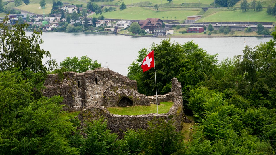 Burgruine mit Schweizerfahne und See im Hintergrund.